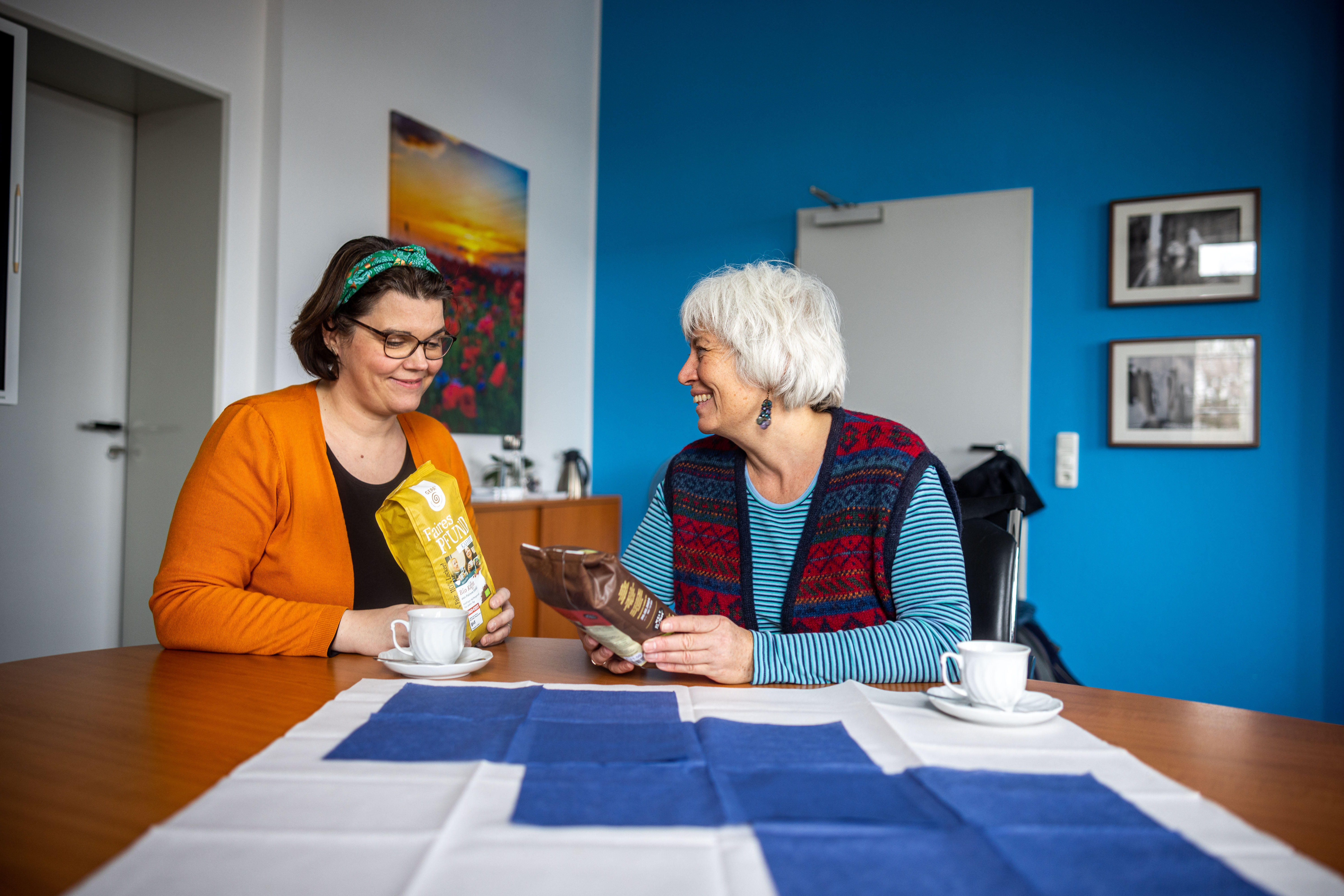 Zwei Frauen sitzen an einem Tisch in einem hellen Raum. Sie lächeln sich an und halten jeweils eine Tüte mit Lebensmitteln. Auf dem Tisch stehen zwei Kaffeetassen und eine Tischdecke mit blauem und weißem Muster.