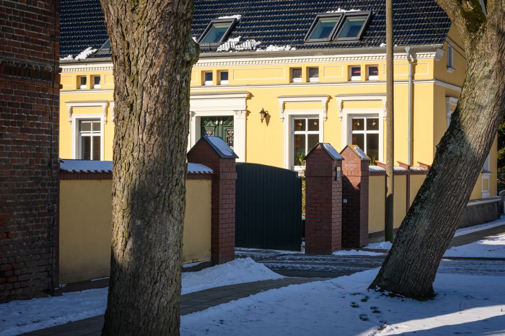 Ein gelbes Gebäude mit weißen Fenstern und einem geschmückten Eingang ist durch eine schwarze Toröffnung sichtbar. Rundherum sind Schnee und Bäume zu sehen, die eine winterliche Atmosphäre schaffen. Der Eingang wird von roten Backsteinpfeilern flankiert.