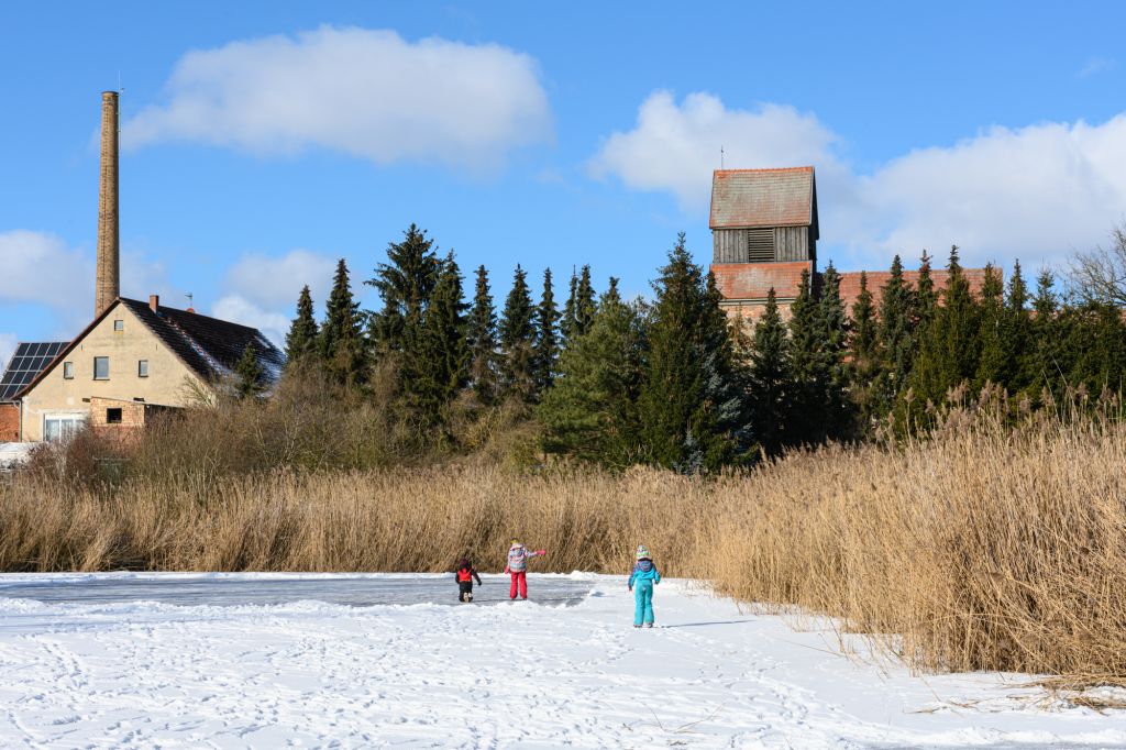 Im Vordergrund spielen Kinder auf verschneitem Boden. Im Hintergrund sind ein Gebäude mit Schornstein und eine Kirche sichtbar, umgeben von Bäumen und hohem Gras. Der Himmel ist blau mit wenigen Wolken.