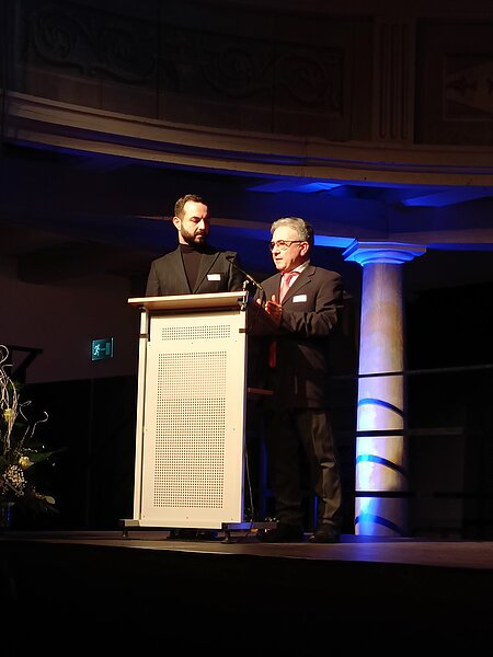 Zwei Männer stehen an einem Rednerpult auf einer Bühne. Der Mann links trägt einen schwarzen Anzug und schaut zur Seite, während der Mann rechts in einem grauen Anzug spricht. Im Hintergrund ist eine festliche Bühnenbeleuchtung sichtbar.