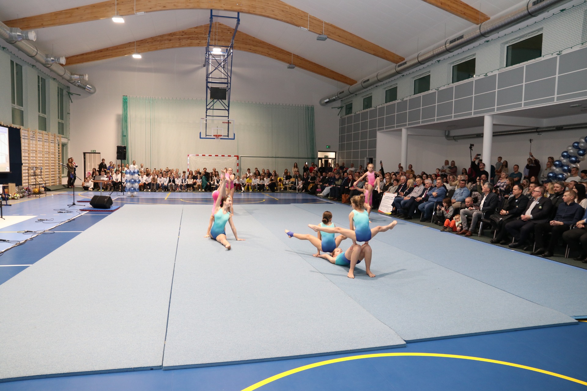 Drei Turnerinnen in bodenlangen Bodenkleidung führen eine synchronisierte Turnübung auf einer blauen Turnmatte vor einem Publikum in einer Sporthalle auf. Im Hintergrund stehen Zuschauer, die die Aufführung beobachten.