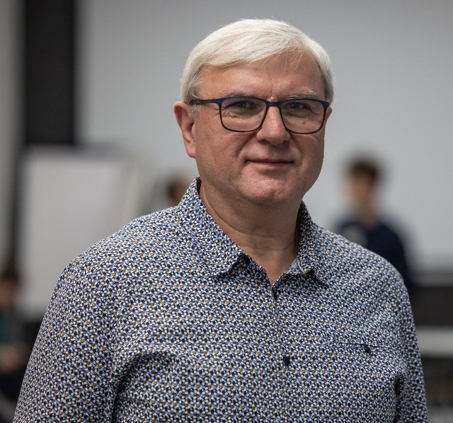 Ein Mann mit kurzen, grauen Haaren und einer Sehhilfe trägt ein gemustertes Hemd. Er steht im Vordergrund, während unscharfe Personen im Hintergrund zu sehen sind. Die Umgebung wirkt wie ein Veranstaltungsraum.