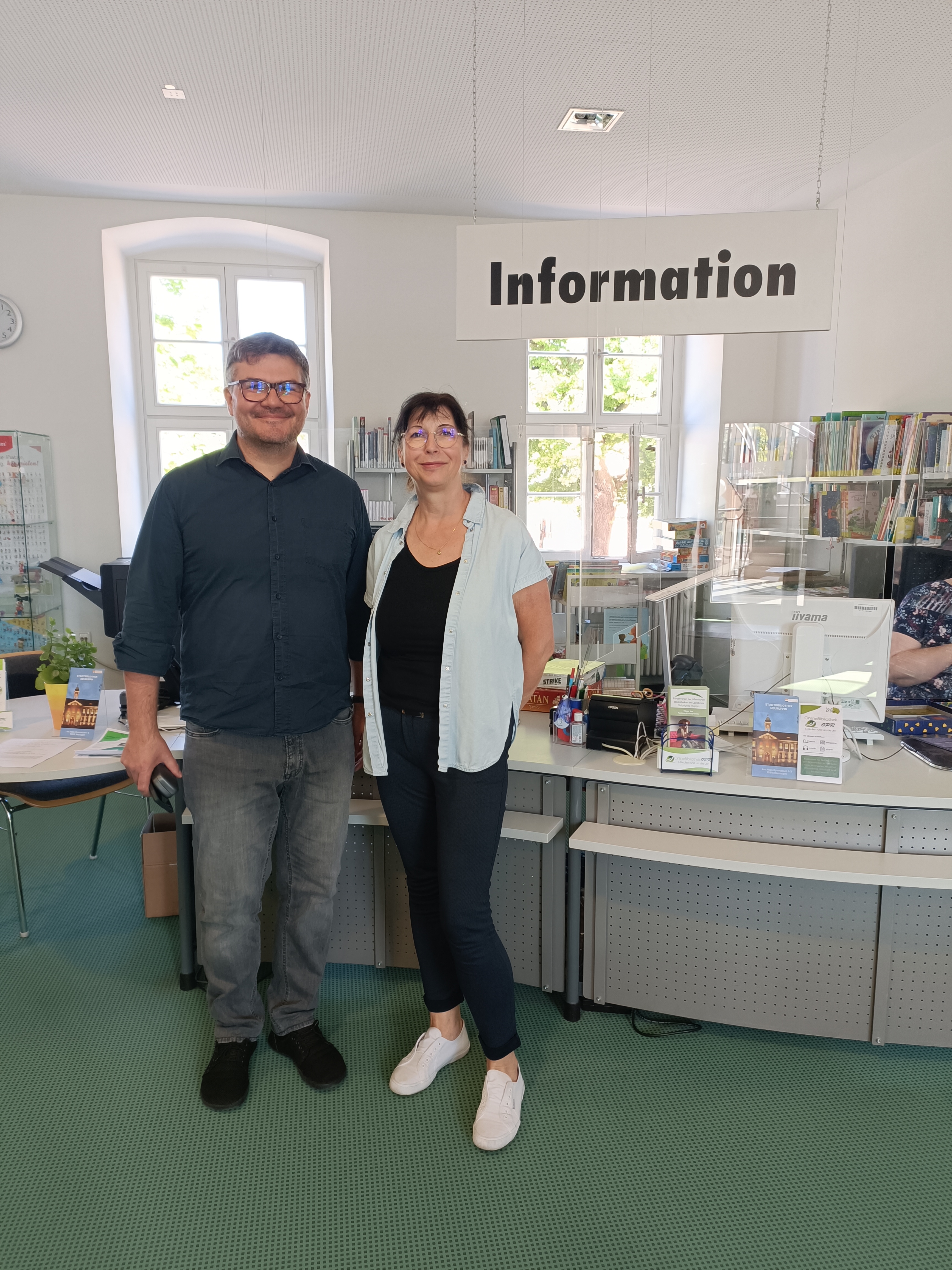 Zwei Personen stehen an einem Empfangstresen in einer Bibliothek. Über ihnen hängt ein Schild mit der Aufschrift „Information“. Der Mann trägt ein schwarzes Hemd und die Frau ein blaues Oberteil. Im Hintergrund sind Regale mit Büchern sichtbar.