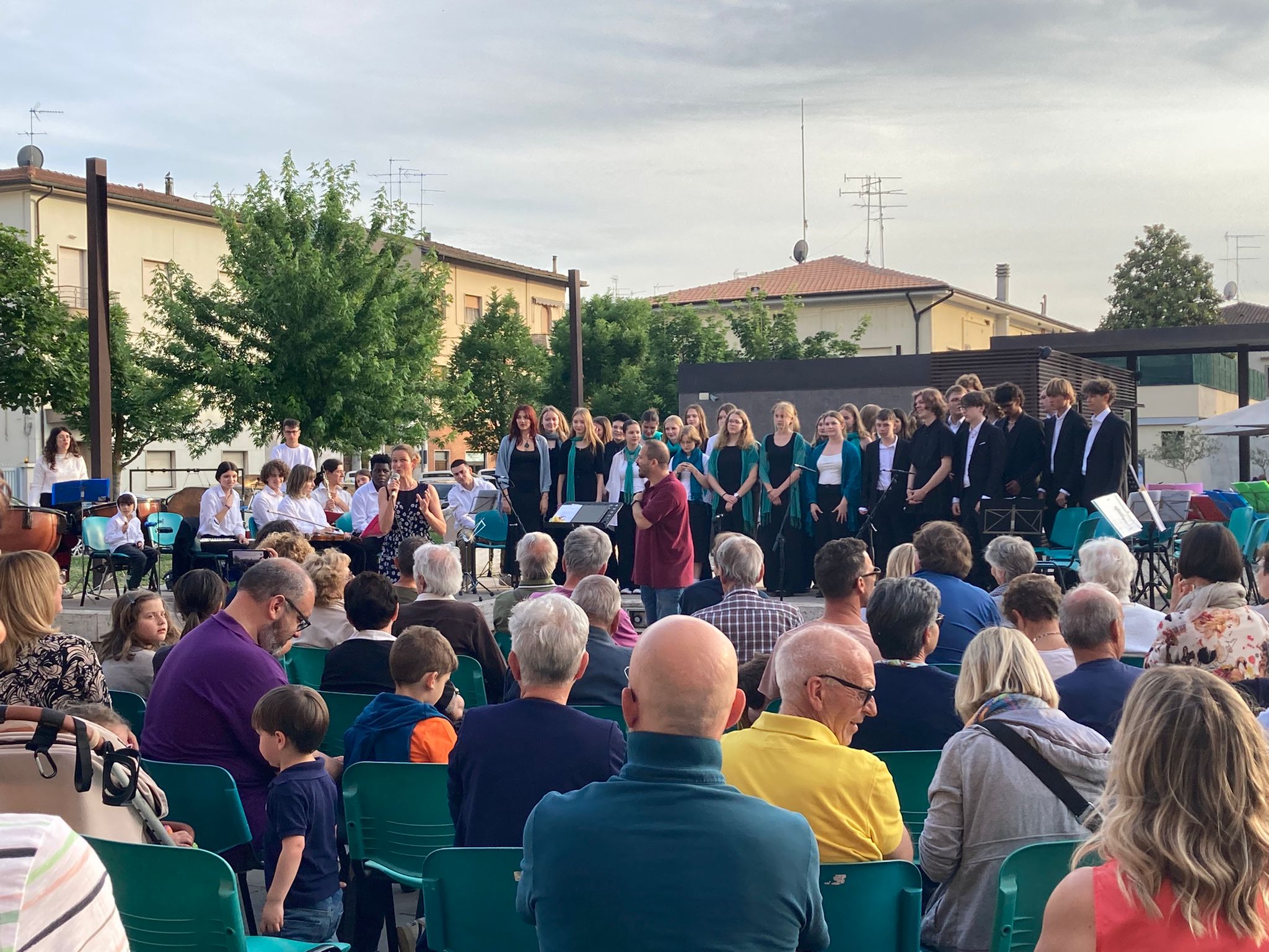 Bei einem Freiluftkonzert stehen eine Gruppe von Sängern und Musikern auf einer Bühne. Die Zuschauer sitzen auf Stühlen und lauschen aufmerksam. Die Atmosphäre ist festlich und gemeinschaftlich, mit Bäumen im Hintergrund.