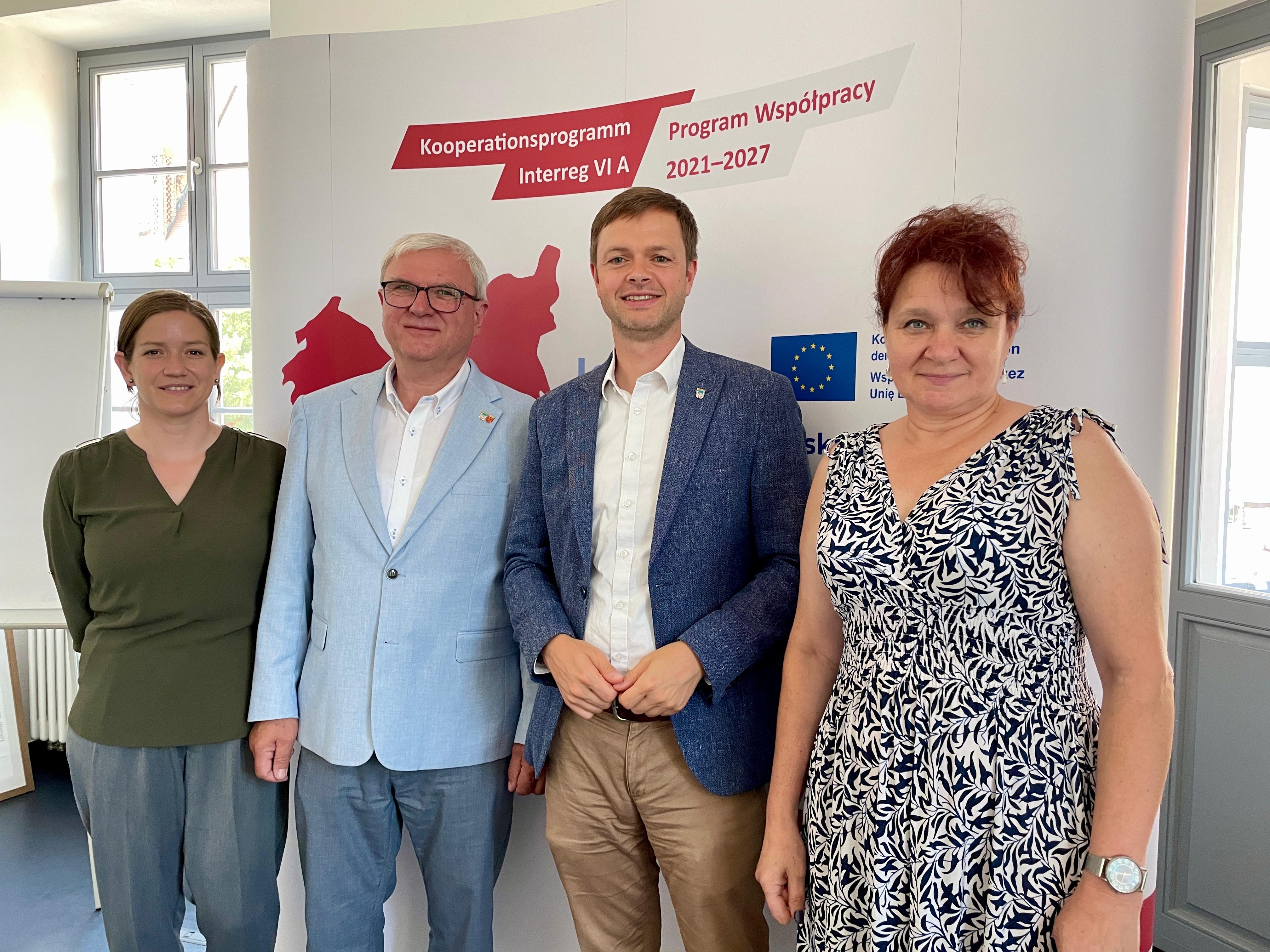 Vier Personen stehen zusammen vor einem Banner, das Informationen zum Kooperationsprogramm Interreg VI A 2021-2027 enthält. Die Personen lächeln und tragen formelle Kleidung. Im Hintergrund sind Fenster zu sehen, die Tageslicht hereinlassen.