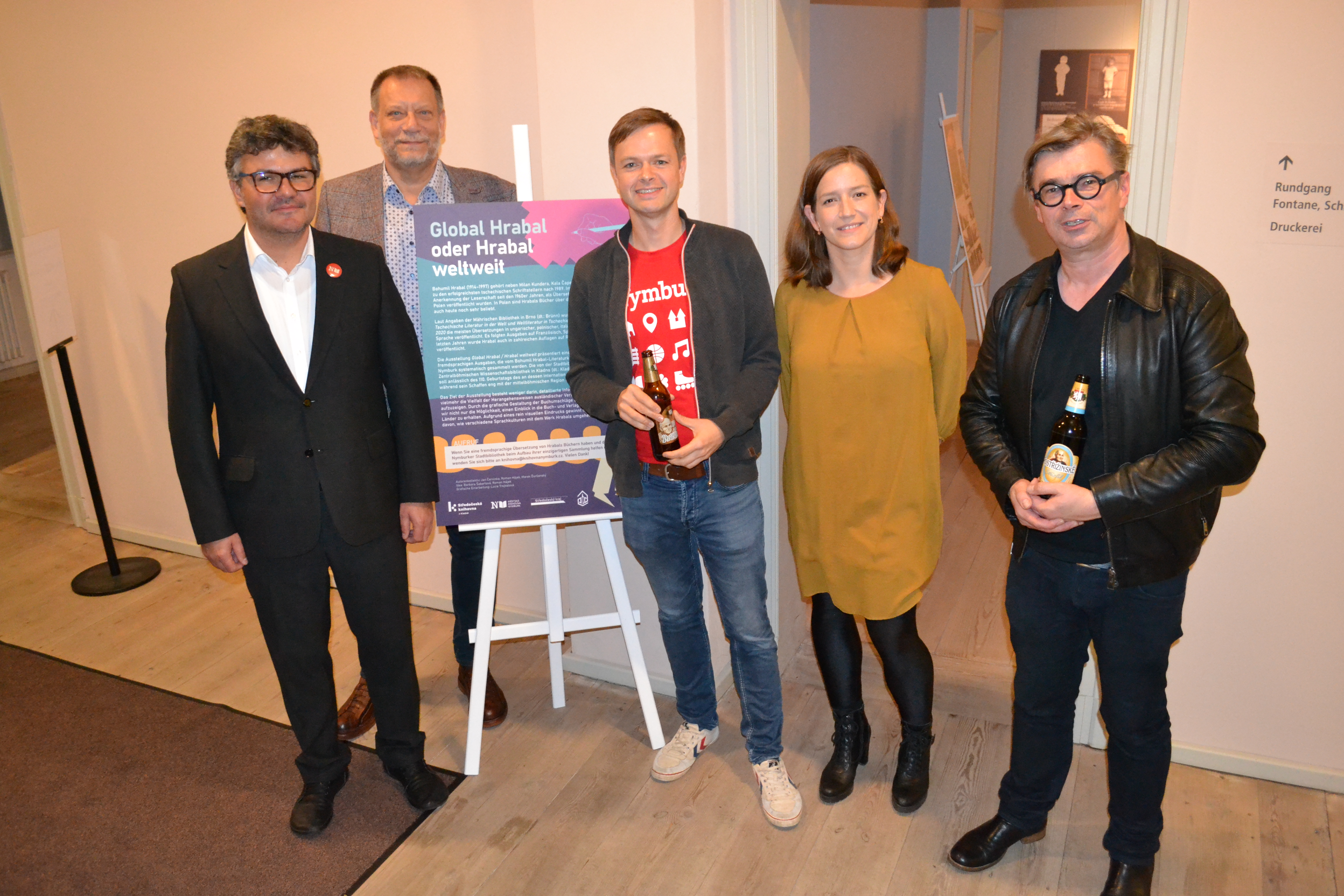 Eine Gruppe von fünf Personen steht vor einem Informationsplakat mit dem Titel „Global Hrabal oder Hrabal weltweit“. Sie lächeln und halten Getränke. Der Raum hat schlichte Wände und einen hellen Boden, Hintergrundelemente sind minimal.