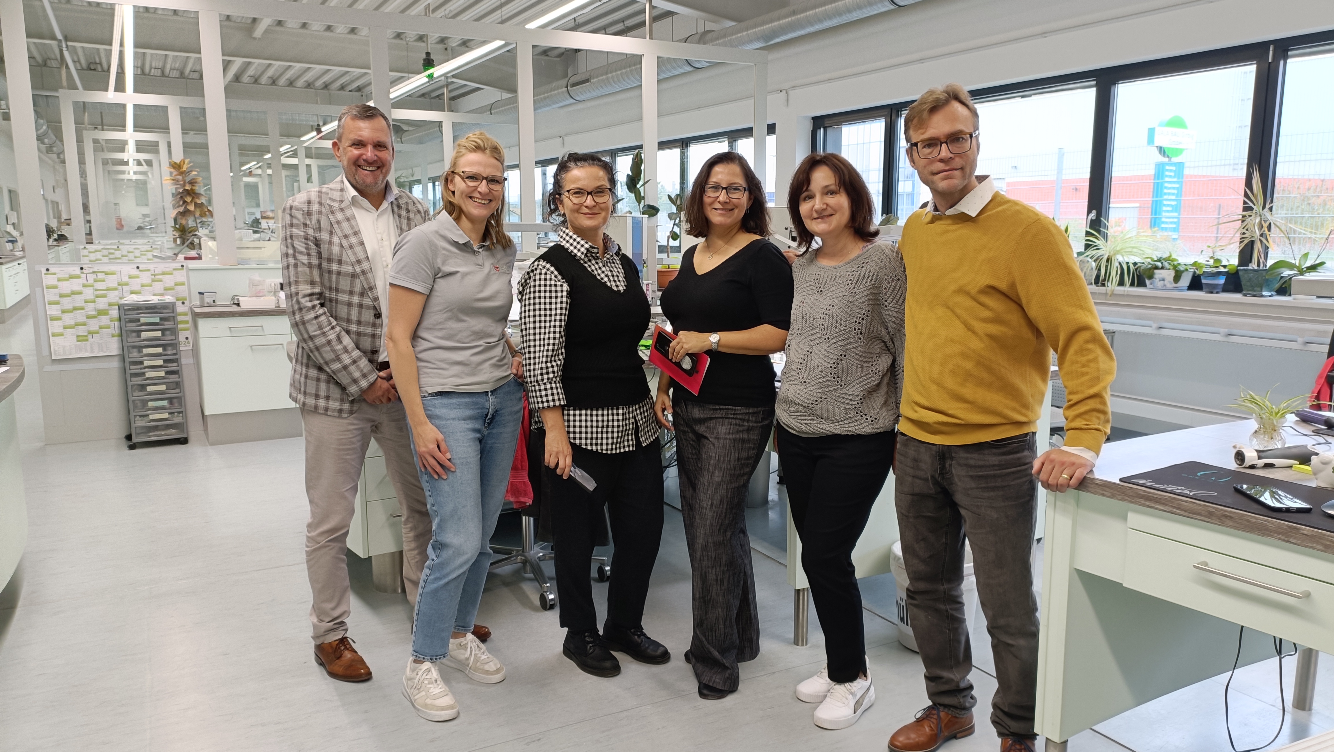 Sechs Personen stehen in einem modernen Büroraum. Sie lächeln und posieren zusammen. Im Hintergrund sind Pflanzen und Büromöbel zu sehen. Die Personen tragen unterschiedliche Kleidung und scheinen ein lockeres, freundliches Umfeld zu genießen.