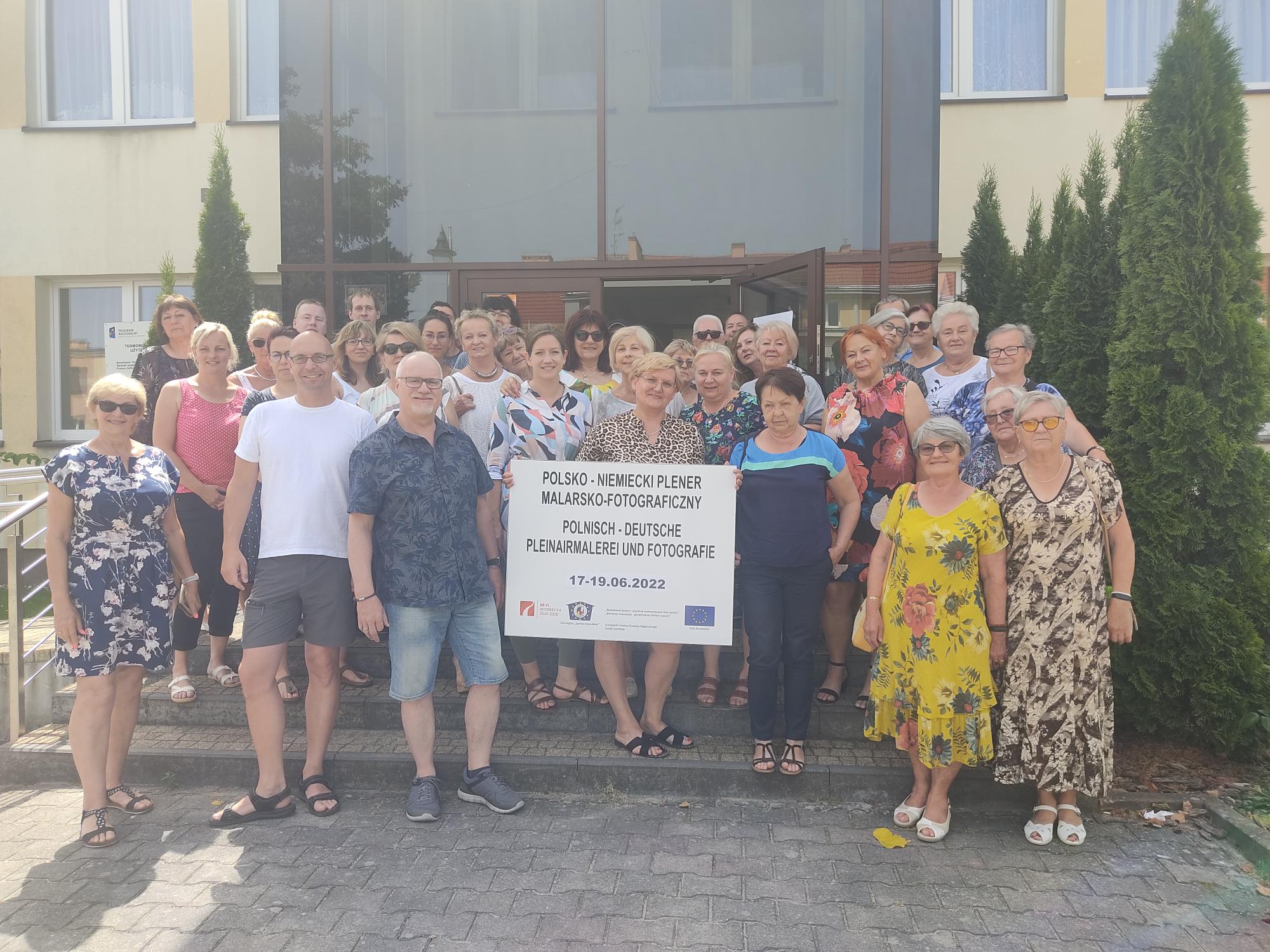 Gruppenfoto vor einem Gebäude: Viele Erwachsene, überwiegend ältere, lächeln in Sommerkleidung. In der Mitte halten Teilnehmende ein großes Schild mit der Aufschrift zu einem polnisch-deutschen Pleinair für Malerei und Fotografie, mit Datum 17.–19.06.2022. Gemeinschaftliches, freundliches Event.
