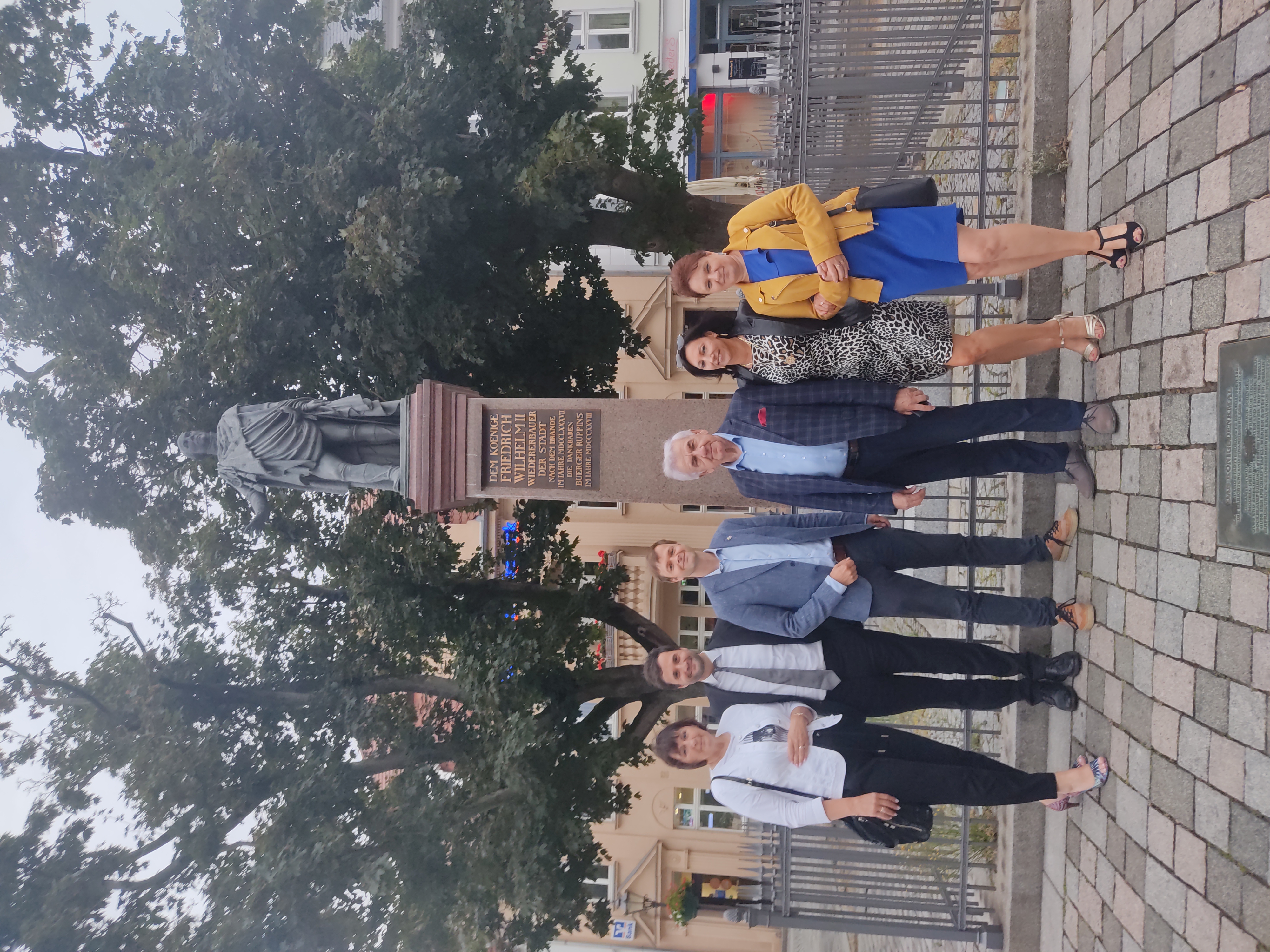 Sechs Erwachsene posieren lächelnd vor einer großen Statue auf einem städtischen Platz. Sie tragen überwiegend elegante Kleidung. Hinter ihnen stehen Bäume und ein umzäunter Sockel der Statue. Die Szene wirkt wie ein Erinnerungsfoto bei einem Besuch oder Termin.