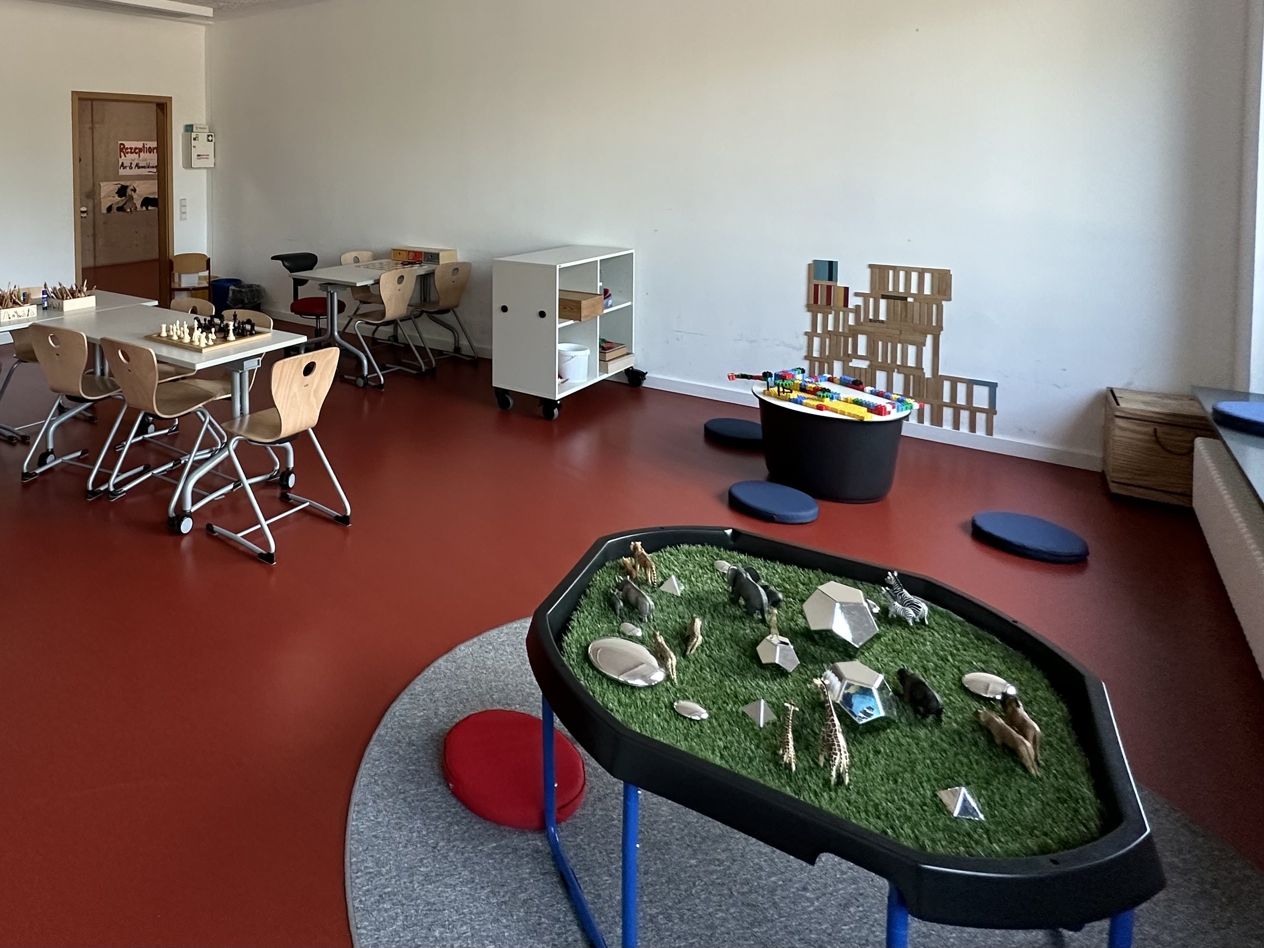 Der Raum zeigt eine Spielsituation mit einem Tisch mit Schachspielen und einem anderen Tisch für Aktivitäten. In der Mitte befindet sich ein Spieltisch mit einer grünen Fläche und verschiedenen Spielobjekten. An der Wand ist eine bunte Spielstruktur aus Holz sichtbar.