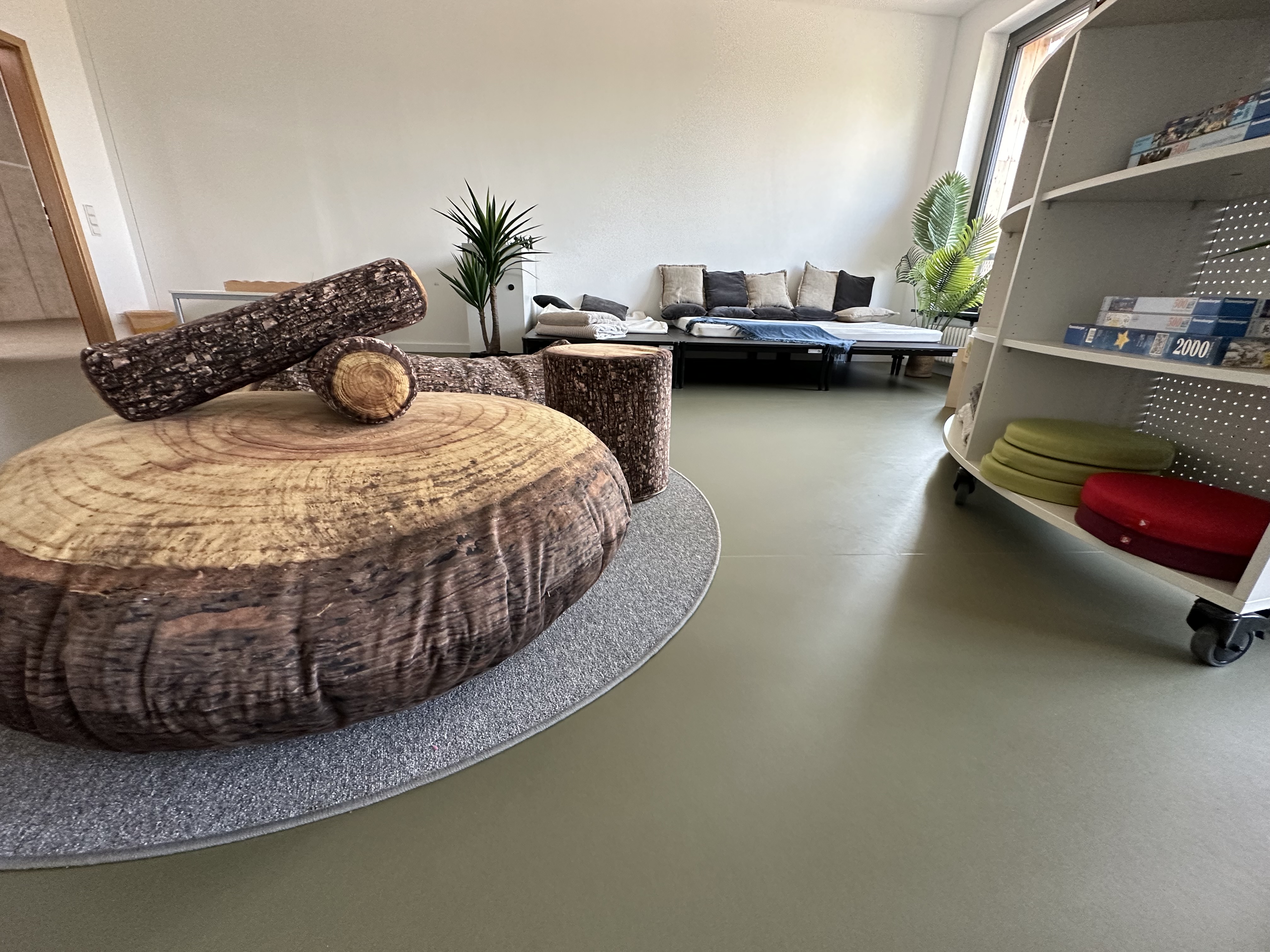 Ein moderner Raum mit einem großen runden Tisch aus Holzscheiben und einem kleinen, hölzernen Zylinder in der Mitte. Im Hintergrund steht ein gemütliches Sofa und Pflanzen sorgen für eine einladende Atmosphäre. Regale mit verschiedenen Materialien sind ebenfalls sichtbar.