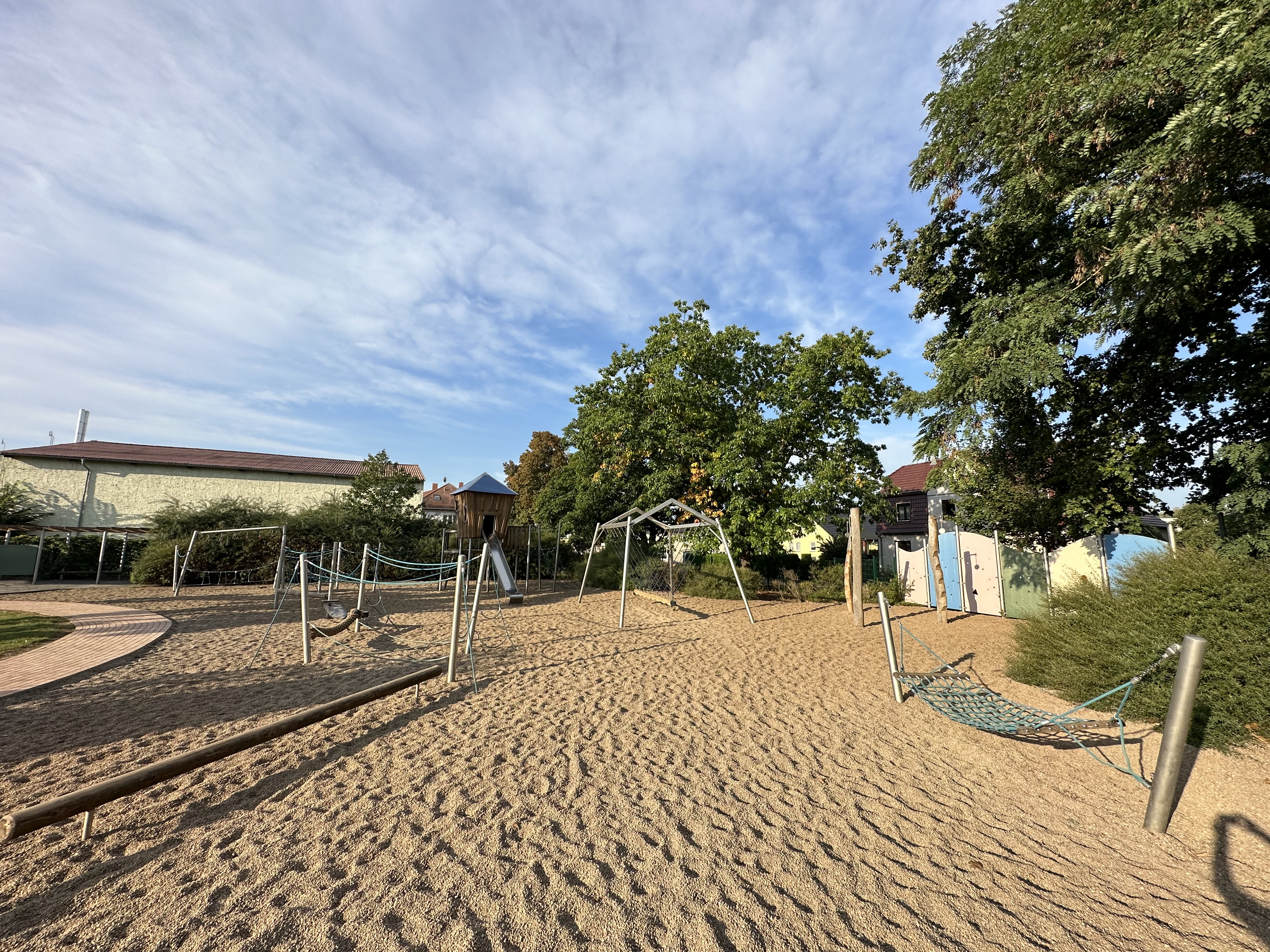 Ein Spielplatz mit Sandboden ist zu sehen. Es gibt Schaukeln, ein Klettergerüst und eine Hängematte. Im Hintergrund sind Bäume und Gebäude sichtbar. Der Himmel ist teilweise bewölkt, was auf gutes Wetter hinweist.