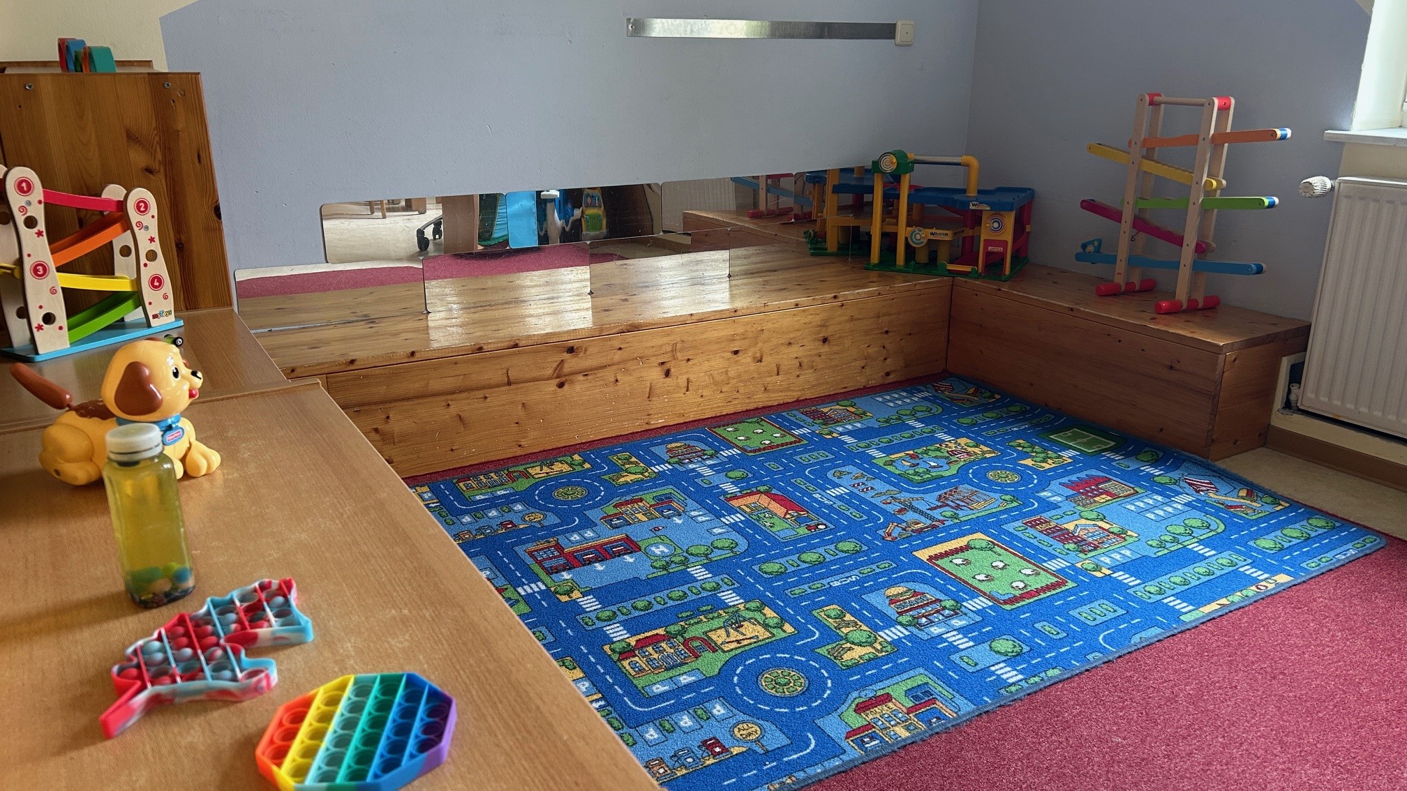 Im Bild ist ein Spielzimmer zu sehen. Der Boden ist mit einem bunten Teppich in Straßen- und Hausdesign gestaltet. Auf einem Tisch liegen einige Spielzeuge, darunter ein Hund und zwei Spielgeräte. An einer Wand befinden sich Regale mit weiteren Spielzeugen.