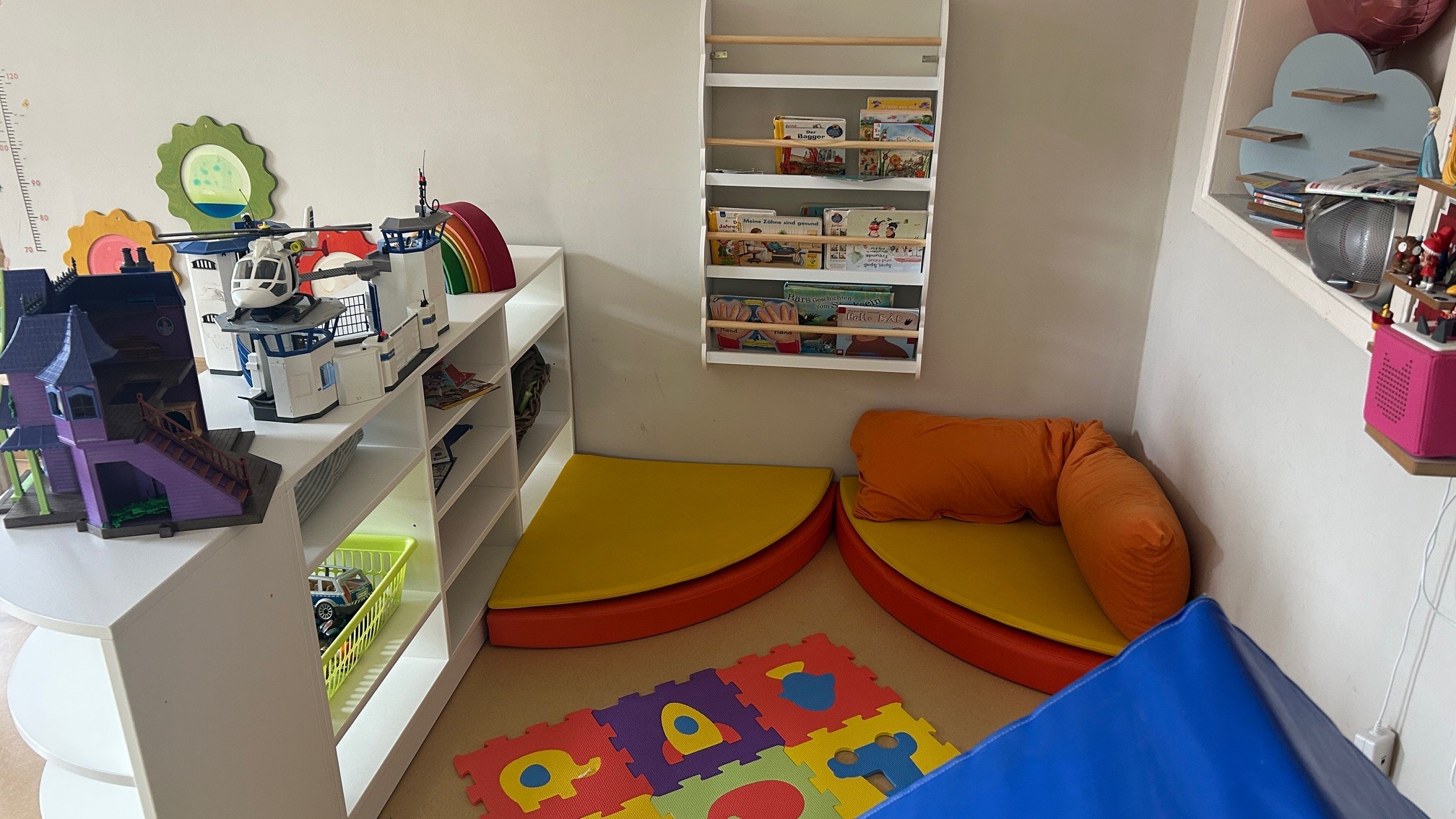 Ein heller Spielraum mit einer Spielecke. Auf einer weißen Regalwand stehen Spielzeug und Bücher. In der Ecke befinden sich zwei gepolsterte Sitzflächen, eine gelb und eine rot. Der Boden ist mit einem bunten Kinderteppich mit Buchstabenmuster ausgestattet.