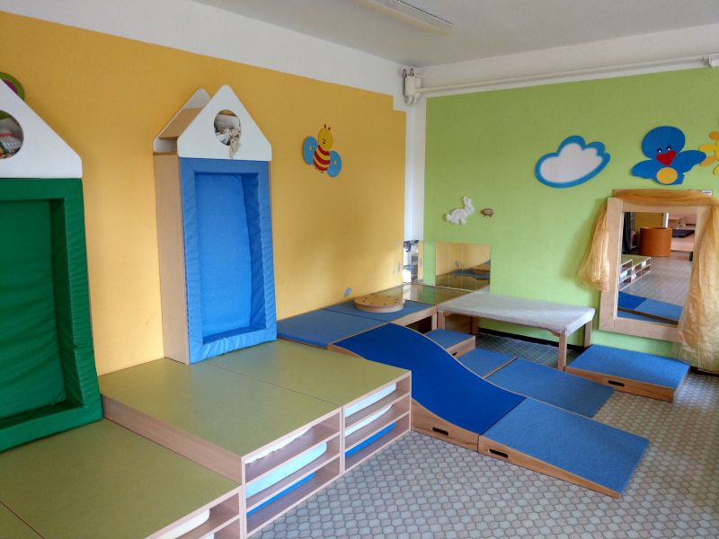 Der Raum ist mit bunten weichen Spielelementen ausgestattet, darunter zwei Klettergerüste in Grün und Blau. Auf dem Boden liegen gepolsterte Matten und ein Steg. An den Wänden sind freundliche Tier- und Wolkenmotive zu sehen, die eine einladende Atmosphäre schaffen.