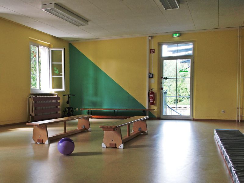 Ein heller Raum mit gelben Wänden und einem grünen Akzent. Auf dem Boden stehen zwei Turnbänke sowie ein lila Ball. Ein offenes Fenster lässt Tageslicht herein, während die Tür zum Außenbereich sichtbar ist.