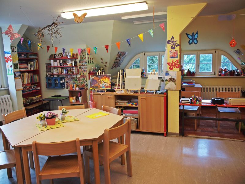 Ein helles Klassenzimmer mit einem runden Tisch und Stühlen in der Mitte. Bunte Girlanden und Schmetterlinge schmücken die Wände. An einer Wand stehen Regale mit Büchern und Materialien, während auf einer anderen Seite Maleasel und Bastelmaterialien bereitliegen.