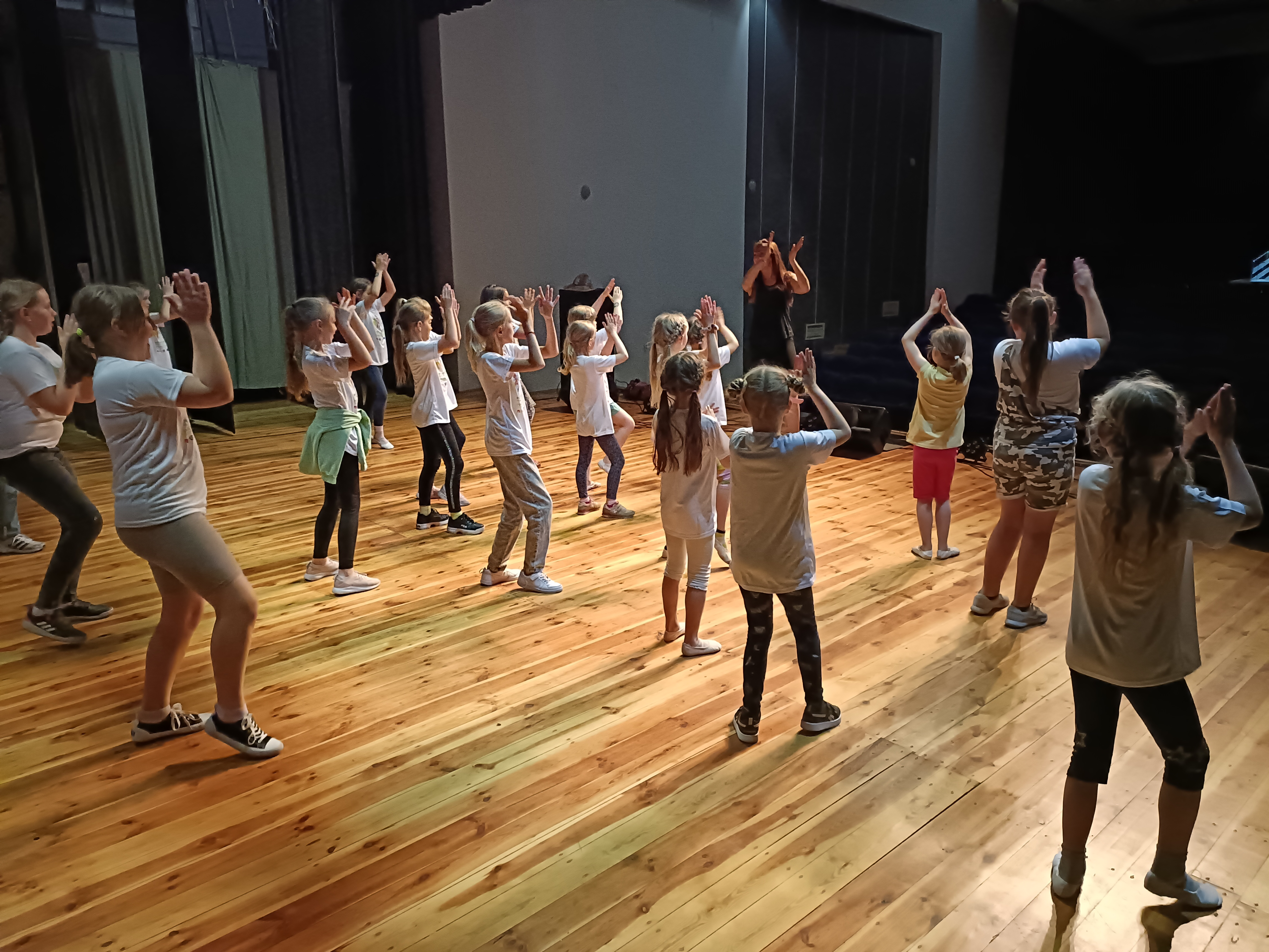 Gruppe von Kindern übt gemeinsam eine Tanzchoreografie auf einer Bühne. Sie tragen Freizeitkleidung und folgen einer Trainerin vorn, Arme hoch, in Bewegung. Holzboden, Bühnenlicht; Fokus auf gemeinsames Training und Rhythmus.