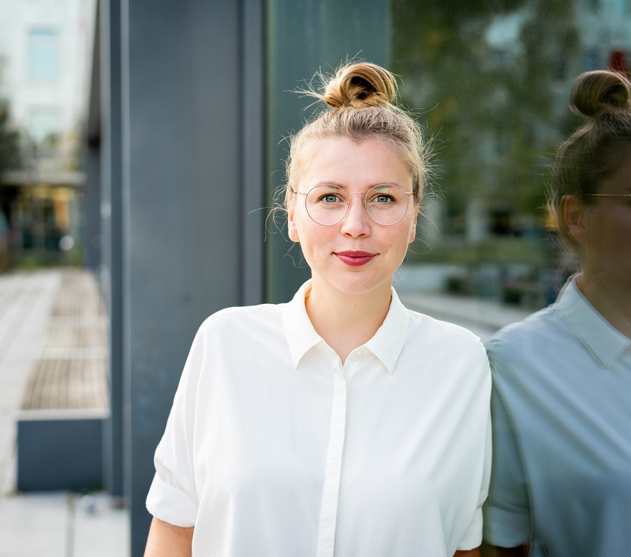 Eine junge Frau mit blonden, hochgesteckten Haaren und einer weißen Bluse steht vor einem Fenster. Sie trägt runde Brillen und lächelt freundlich. Im Hintergrund sind unscharfe Bäume und Gebäude zu erkennen.