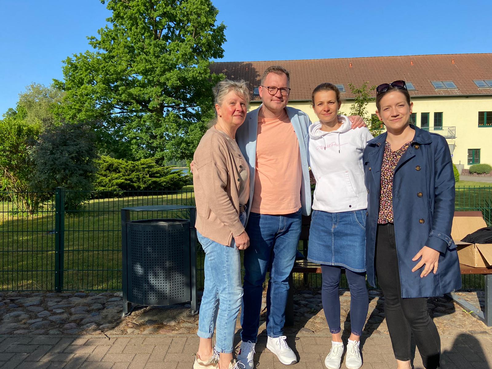 Vier Erwachsene stehen dicht nebeneinander im Freien und lächeln in die Kamera. Der Mann und drei Frauen tragen Freizeitkleidung; zwei haben die Arme umeinander gelegt. Im Hintergrund ein Zaun, Bäume und ein helles Gebäude bei Sonnenschein.