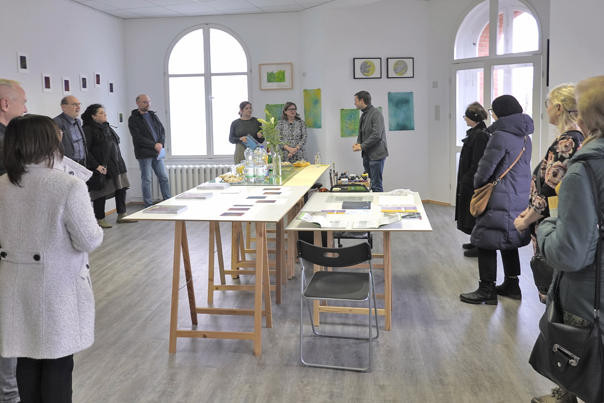 Kleine Gruppe in einem hellen Ausstellungsraum. Menschen stehen im Halbkreis um einen langen Tisch mit Zeichnungen und Materialien. Eine Person spricht und präsentiert die Arbeiten. Bilder hängen an den Wänden; Atmosphäre wirkt wie Vernissage oder Workshop.