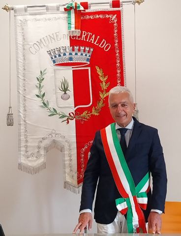 Älterer Mann im dunklen Anzug trägt eine Schärpe in den italienischen Farben. Er steht lächelnd vor einem rot-weißen Gemeindebanner mit Wappen und der Aufschrift „Comune di Certaldo“.